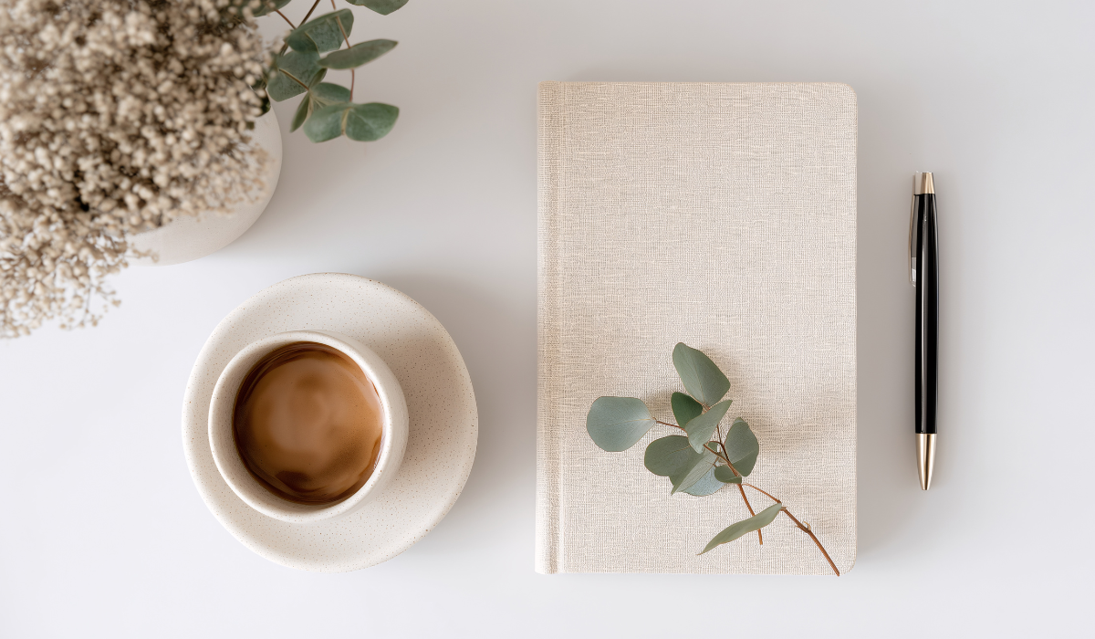 ベージュのノートと黒いペン、コーヒーカップを並べたデスクのフラットレイ